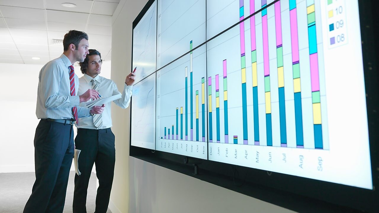Business men analyse reports displayed on a large digital screen