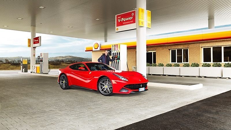 Një Ferrari i kuq i ulur në një stacion të ndërtesës Shell me një burrë të mbështetur në një pompë benzine