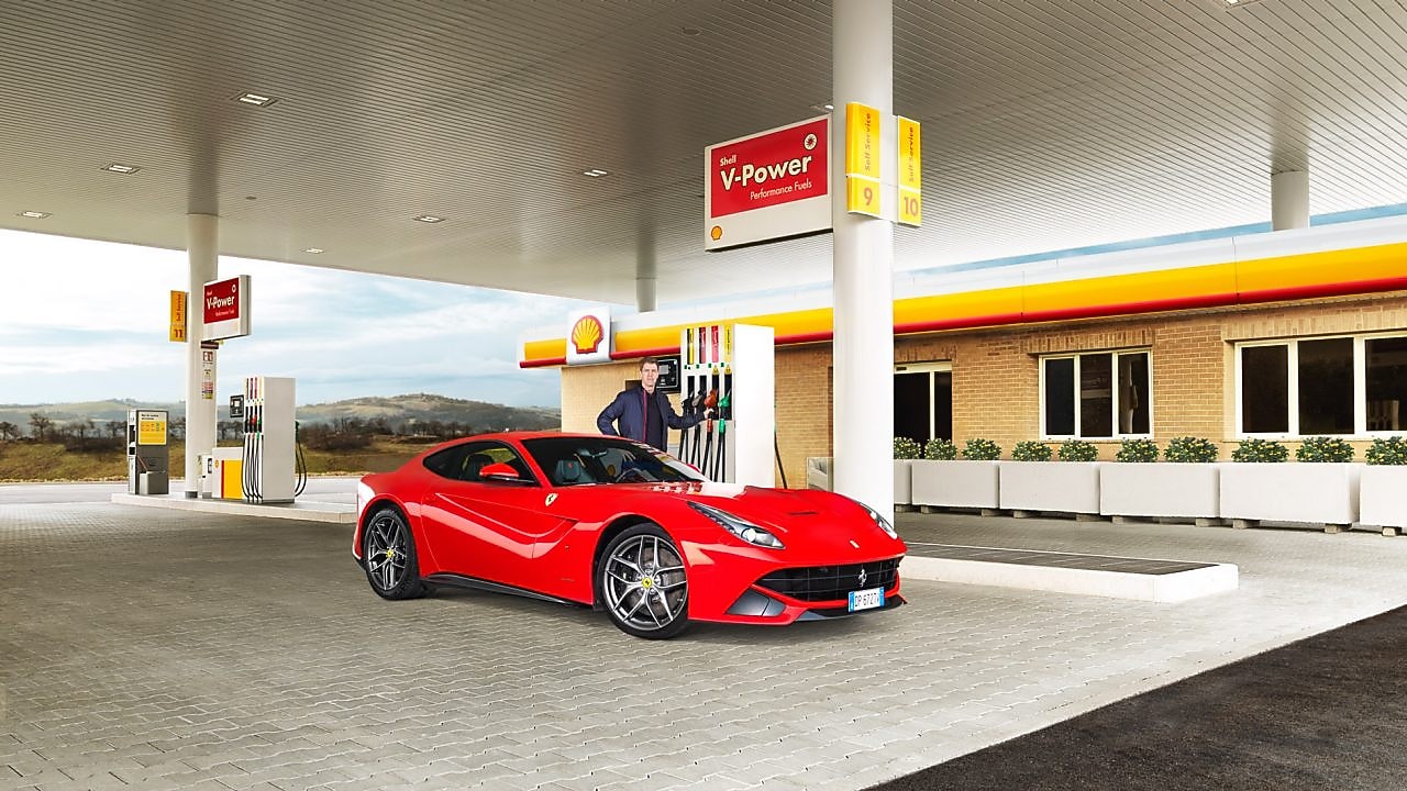 Një Ferrari i kuq i ulur në një stacion të ndërtesës Shell me një burrë të mbështetur në një pompë benzine
